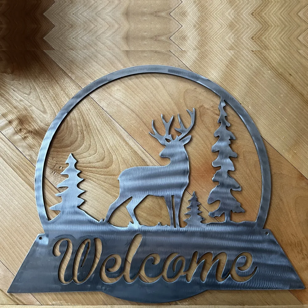 Raw steel Welcome sign with a buck between pine trees.