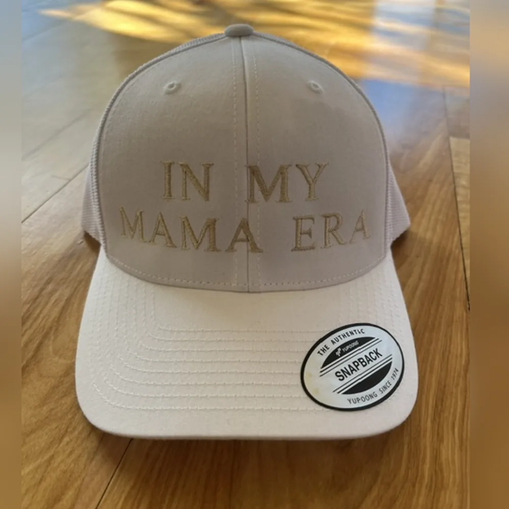 White snapback trucker hat with IN MY MAMA ERA embroidered in tan thread.