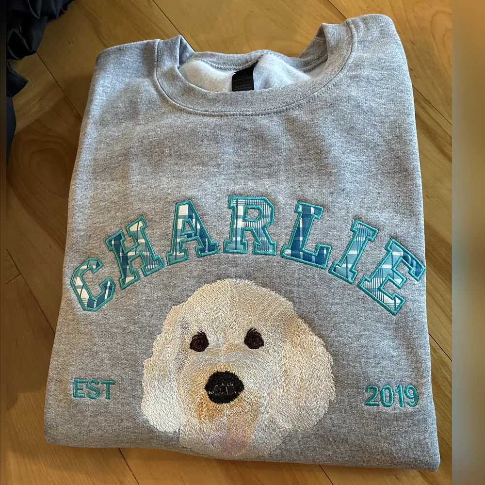 Grey crewneck sweatshirt with CHARLIE in plaid appliqué letters over an embroidered portrait of a white doodle, with EST 2019.