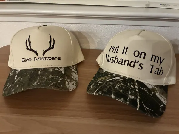 Two cream camo-brim hats embroidered with Size Matters antlers and Put it on my Husband's Tab